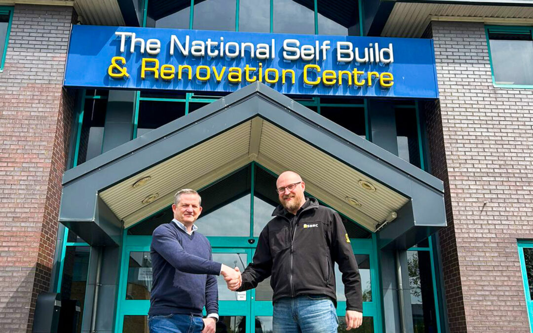A photo of two men shaking hands in front of a large sign that reads "The National Self Build & Renovation Centre." The man on the left is smiling and wearing a Korniche jacket (Ian Bousfield, Made For Trade's Marketing Manager). The man on the right is smiling and wearing a fleece with the NSBRC logo (Gareth Nicholas, NSBRC Business Development Manager).