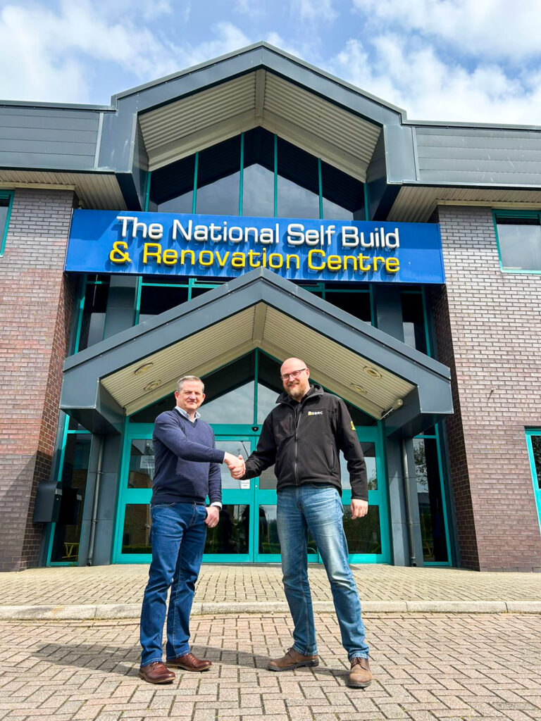 Ian Bousfield, Made For Trade Marketing Manager, and Gareth Nicholas, NSBRC Business Development Manager, shaking hands in front of The National Self Build & Renovation Centre sign.