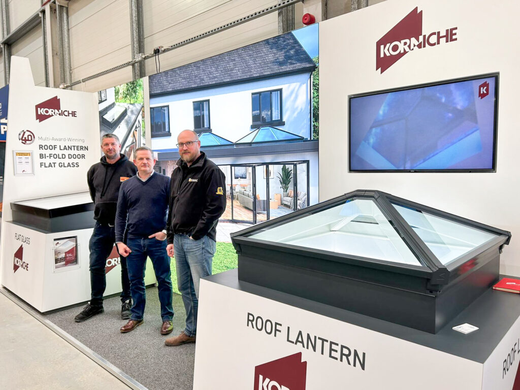 Three people at a Korniche trade show stand. From left to right are: a man in a Sevenday branded hoody (Area Manager from Sevenday Windows, Doors & Conservatories), a man in a jumper smiling (Ian Bousfield, Made For Trade & Korniche Marketing Manager), and a man in a NSRBC branded fleece (Gareth Nicholas, NSBRC Business Development Manager)