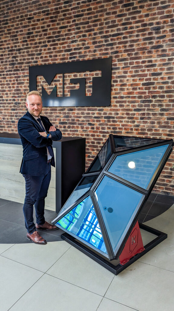 Bradley Gaunt, Managing Director of Made For Trade, in a suit with arms crossed, standing next to a Korniche Roof Lantern with the Made For Trade logo on a brick wall.